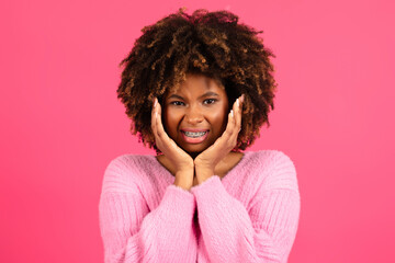 Sad dissatisfied smiling young black curly lady with braces in casual presses hands to cheeks, suffering from toothache