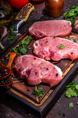 Raw pork steaks cooking background. Three tasty raw fresh meat steaks with herbs, salt, pepper, olive oil, ready for dinner preparation, dark brown kitchen background copy space
