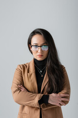 portrait of brunette asian businesswoman in beige blazer and glasses standing with crossed arms isolated on grey