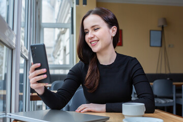 Portrait of smiling young woman using mobile phone while sitting in in coffee shop. White female freelancer connecting to internet on mobile phone.