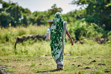 A woman wearing green sari carries pots of food walking through a green village