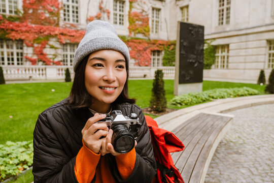 Smiling asian woman taking pictures with vintage camera while sitting on bench in old city