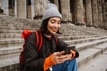 Positive asian woman tourist using mobile phone while sitting on old museum stairs - obrazy, fototapety, plakaty