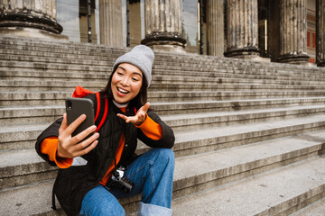 Smiling asian woman making video call via smartphone and gesturing while sitting on stairs