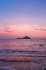 Fototapeta na wymiar Pink color sunset on the ocean with island in background. Concept of travel and scenic place destination. Long exposure on ocean waves. Empty sky on horizon to write your text. Copy space.