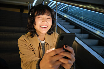 Plakat Close up portrait of smiling asian girl student, listens music in headphones and looks at mobile phone, uses smartphone, sits on staircase in public place
