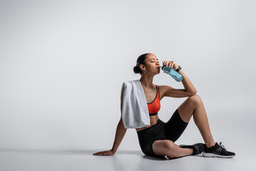 young african american woman in sports bra and bike shorts sitting with towel and drinking water on grey background.