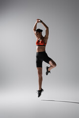 full length of tattooed african american woman in sports bra and shorts jumping with raised hands on grey.