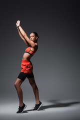 full length of tattooed african american sportswoman in sneakers standing with raised arms on grey.