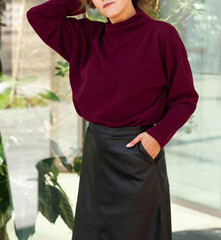 Young blond sexy girl woman in a romantic look in a red marsala winter cosy sweater and black eco leather skirt a in an urban city center new the glass wall with plants.