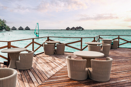 A wide-angle shot of an empty street bar with multiple wicker tables and chairs placed on a wooden pier with a sail, swimming people, and a group of hotel bungalows in a defocused background; Maldives