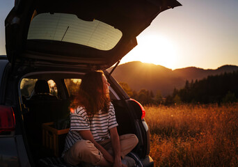 Young cheerful woman traveler sitting in open trunk of car enjoying sunset inmountains during solo...