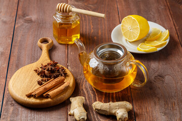 Transparent teapot of lemongrass tea next to anise, ginger and cinnamon on a round wooden board and honey and lemon.