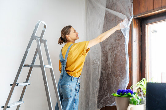 Young adult happy woman renovating the house, standing on stepladder covering the wall with film before painting