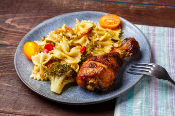 Pasta with vegetables and cherry tomatoes with baked chicken leg on the table next to a light napkin and fork.
