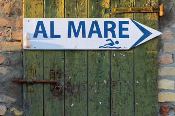 Old wooden door and direction arrow sign pointing to the right with the italian language words Al Mare meaning To the Sea, with graphic shape of a man swimming - 566330170