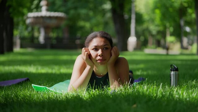 Thoughtful African American woman lying on yoga mat in park, waiting for coach