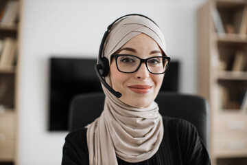 Smiling positive confident woman in hijab and headset sitting at office desk. Company muslim worker looking at camera. Concept of people and business.