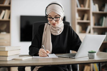 Attractive confident muslim business woman, office manager, wearing headset and hijab using laptop...