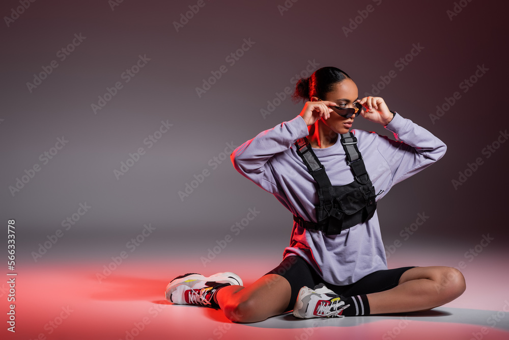 Wall mural full length of african american woman in bike shorts and sweatshirt wearing sunglasses on grey background with red light.