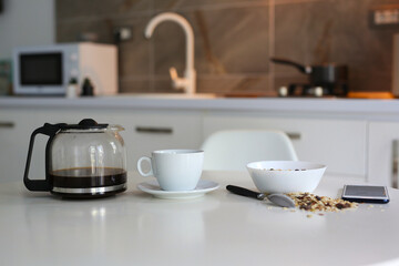 morning in a cozy apartment, on a white table a kettle with fresh black coffee, a white cup and muesli in a plate, a mobile phone lies nearby