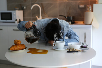 a beautiful girl in a denim shirt, her head rested on the table, she sleeps and pours coffee past the mug, the girl did not get enough sleep