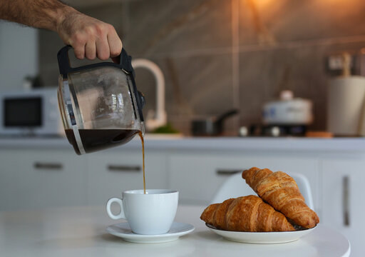 a man holds a teapot with coffee and pours into a white cup, a hot fresh drink, a plate of croissants stands nearby, all this against the backdrop of a beautiful white kitchen