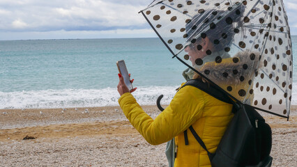 Mujer joven con paraguas en la playa haciendo una videollamada debajo de un paraguas en un día lluvioso