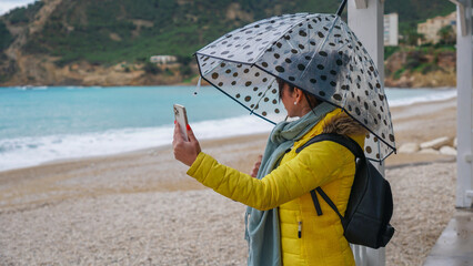 Mujer joven con paraguas en la playa haciendo una videollamada debajo de un paraguas en un día lluvioso