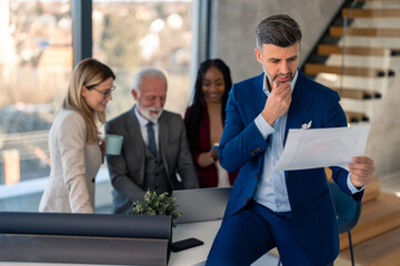 Concentrated entrepreneur male manager analyzing paper document developing business strategy. Diverse multiethnic corporate team brainstorming working on laptop in background.