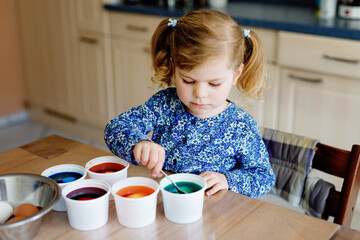 Excited little toddler girl coloring eggs for Easter. Cute happy child looking surprised at colorful colored eggs, celebrating holiday with family. Adorable kid at home with different bright colors.