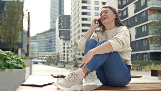 Young cheerful woman talking on a smartphone while sitting on the street of a modern European city.