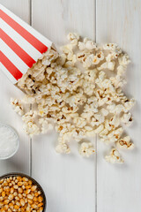popcorn bag, popcorn kernels and salt on a white wooden table, top view.