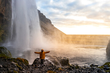 Girl in yellow raincoat with arms outstretched stands close to stunning powerful famous Seljalandsfoss waterfall, southern, Iceland