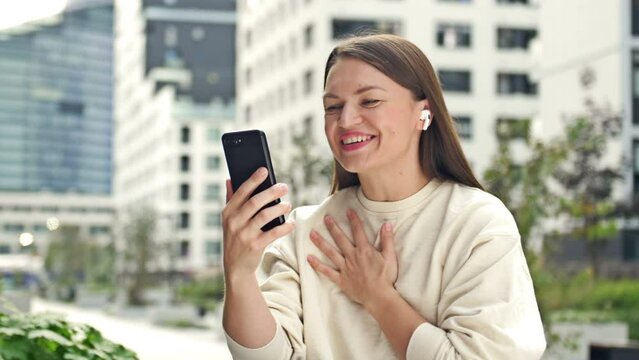 Young cheerful woman with an earphone in her ear is talking on a smartphone while sitting on the street of a modern European city.