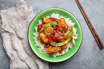 Asian sweet and sour sticky chicken with vegetables stir-fry and rice in ceramic bowl with chopsticks top view on gray rustic stone background, traditional Asian dish