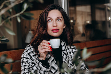 A beautiful brunette woman in a hounds tooth check coat sits in an outdoor cafe and drinks coffee