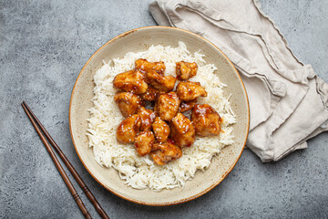 Chinese sweet and sour sticky chicken with sesame seeds and rice on ceramic plate with chopsticks top view on gray rustic stone background, traditional dish of China