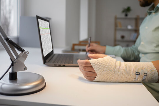 Bearded man with bandaged broken arm is working at laptop, sitting at his desk and making notes in notebook for reporting. Recovery from hand injury. Social security and health care concept.