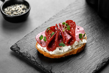 Sandwich, toast with sliced salami, pepperoni, cream cheese, sausage, microgreen on dark slate cutting board. Snack, bruschetta.