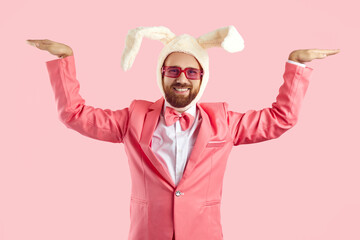 Smiling funny man in suit and bunny ears isolated on pink studio background show moves. Happy guy...