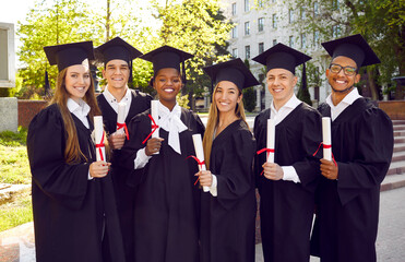 Group portrait of happy diverse university graduates in green campus yard on graduation day. Six cheerful joyful multiracial students in caps and gowns standing together, looking at camera and smiling