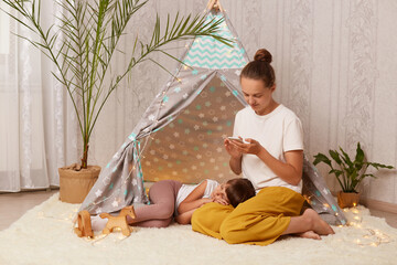 Photo of Caucasian brown haired woman sitting on floor near children's wigwam and using cell phone while her daughter sleeping on her legs, female reading news, browsing internet. © sementsova321