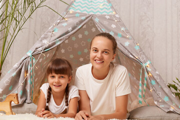 Image of pretty positive pleased woman lying on floor near children's wigwam with her charming daughter, looking at camera, playing together in kid's bedroom, expressing happiness. © sementsova321