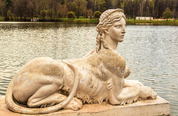 An ancient park sculpture in the form of a female lion sphinx on the shore of a pond in the park