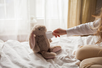 Curly-haired little blue-eyed girl of 4 plays with her plush rabbit in tea party. Children's room....