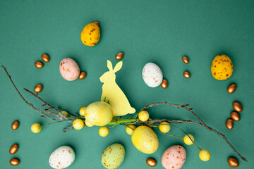 Chocolate colorful decorative eggs and decorative branch with rabbit on a green background. Happy Easter background. Flat lay