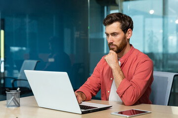 A young businessman, freelancer, designer in a red shirt sits in the office at the table and works on a laptop. He looks thoughtfully at the monitor, holds his chin with his hand.