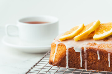 Loaf of orange bread covered with a confectionery glaze with lemon juice and decorated with orange slices. Chiffon cake on a pastry grill next to cup of tea. White background