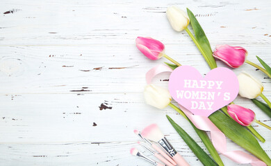 Flowers of tulips, card in shape of heart with text Happy Women's Day, pink makeup brushes and ribbon on white wooden background